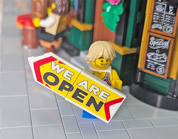 LEGO minifig holding a custom made and printed 'WE ARE OPEN' sign in front of a pub made from lego bricks. LEGO MOC